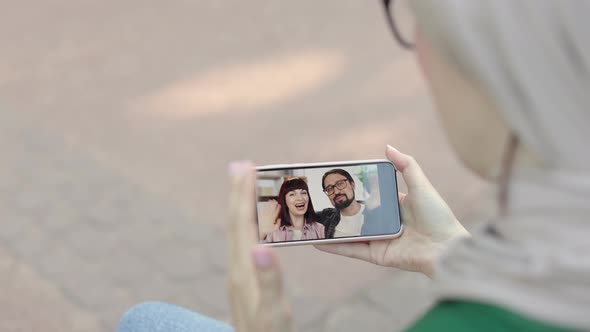 Lady in Stylish Hijab Sitting on the Bench at Park and Talking Online with Her Friends alt