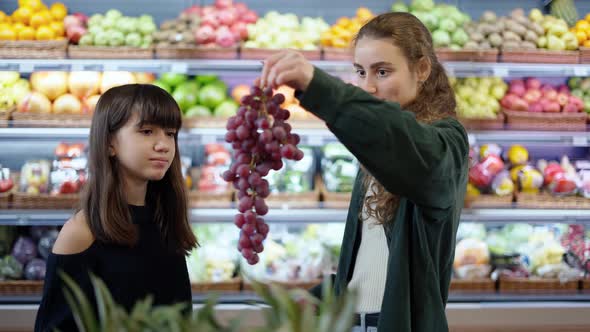 Two Cheerful Girls Choosing Bio Fruits  Grape in Supermarket Together alt