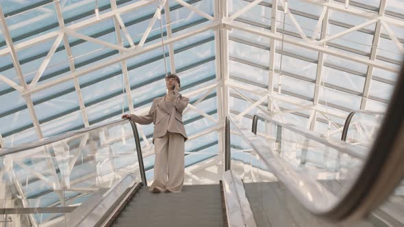 Young Businesswoman Going down Escalator and Talking on Smartphone alt