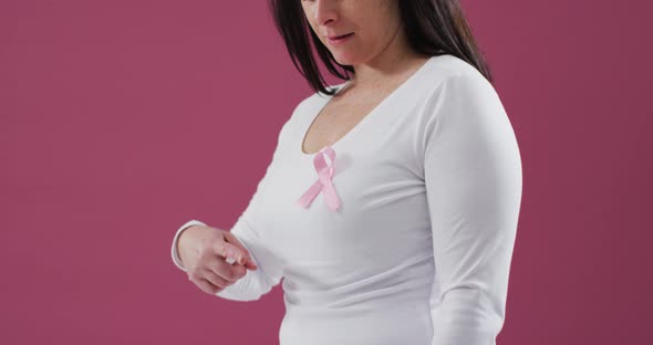 Mid section of a woman pointing to the pink ribbon on her chest against pink background alt
