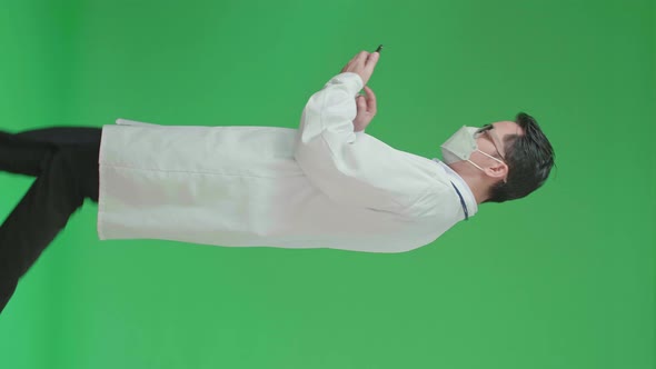 Asian Doctor With Stethoscope Wearing Mask, Using Mobile Phone While Walking On Green Screen alt