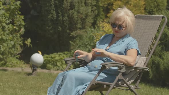 Carefree Female Retiree Sitting in Garden Chair and Applying Hand Moisturizer. Portrait of Happy alt