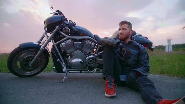 Middleaged Biker with Beard Sits By Motorcycle at Sunset