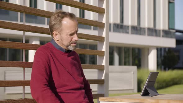 A Middleaged Handsome Caucasian Man Looks at a Tablet and Smiles As He Sits at a Table in a Park alt