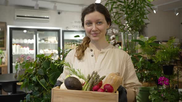 Smiling Woman Holds Box of Vegetables and Looks at Camera alt
