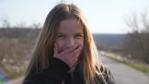 Close Up of Happy Little Girl Looks Into Camera Outdoor and Laughs Covering Her Face with Hand alt