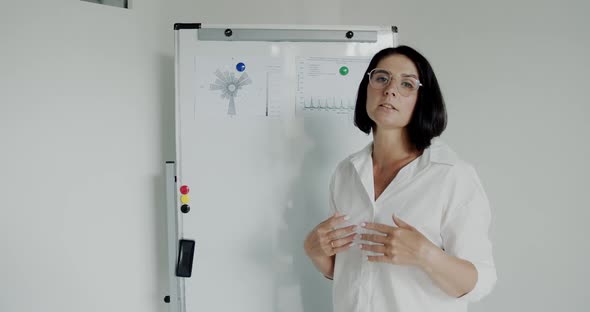 Female Speaker Explaining Topic with Gesturing in Front of Camera at Whiteboard alt