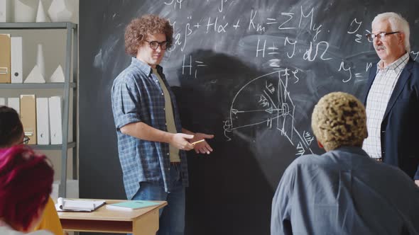 Male High School Student Answering Questions of Professor beside Chalkboard alt