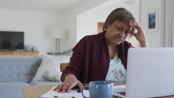 Worried african american senior woman at dining table, using laptop and holding head alt