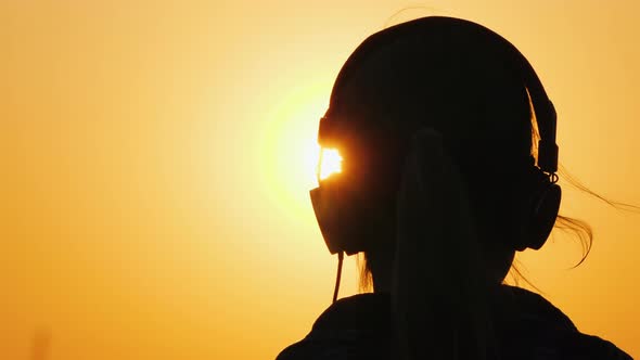 Rear View of a Child with Headphones Listening To Music and Admiring the Sunset and a Big Orange Sky alt