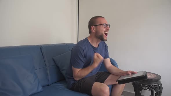 Man wearing eye glasses sitting on the sofa, turns the tablet on and sees a positive outcome. alt