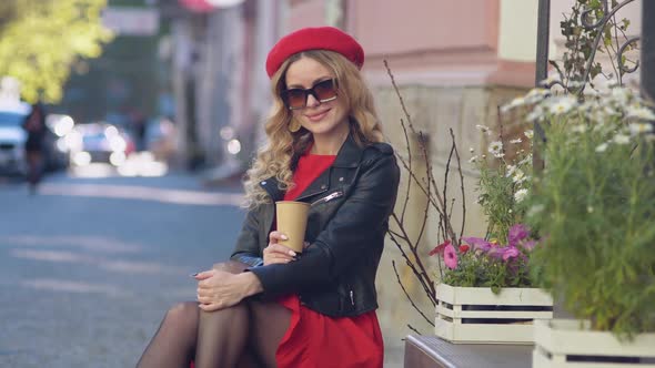 Young Beautiful Woman in a Red Beret and Dress Drinking Coffee and Looks at the Camera alt