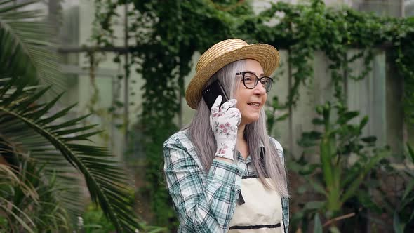Smiling Grey-Haired Woman-Pensioner in Hat Walking in Greenhouse and Talking on Phone alt