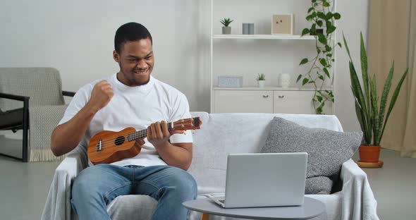 Afro American Guy Student Wears Casual Clothes Sitting on Sofa with Musical Instrument Playing alt