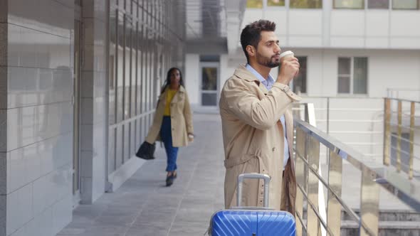 Confident Businessman Traveler Standing Outside Modern Building with Suitcase Drinking Coffee alt