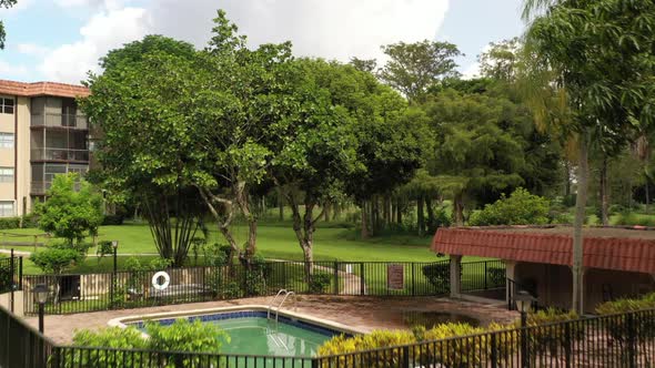 An aerial view over an empty pool, surrounded by green grass and trees on a sunny day in an apartmen alt