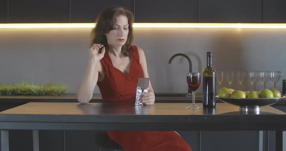 Gorgeous Depressed Mid-adult Woman in Red Dress Sitting in Kitchen and Eating Chocolate. Portrait of alt
