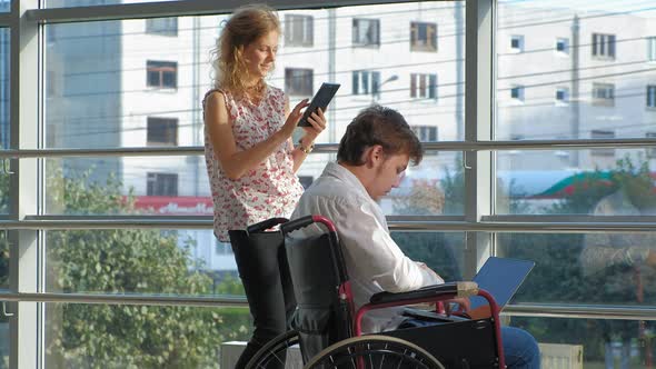 Disabled Businessman on a Wheelchair at a Window with a Laptop Near Woman Uses a Tablet