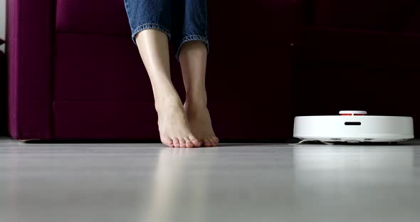 robot vacuum cleaner driving around barefoot woman at home. Female lift feet up when a smart vacuum 