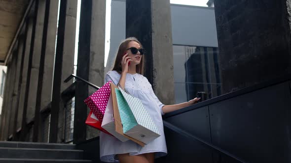 Girl Staying Near Shopping Mall with Shopping Bags and Talking on Mobile Phone About Purchases alt