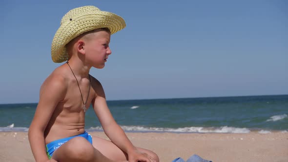 Boy Sits on the Golden Sand. The Child Wears a Straw Hat with a Large Brim alt