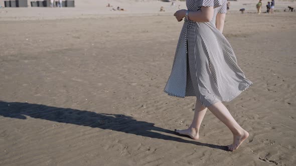 A Woman in a Flowing Dress Walks on an Ocean Beach on a Windy Sunny Day