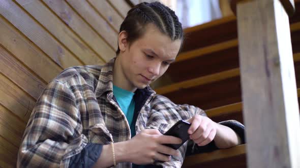 Teen Boy Sitting on Stairs at Home and Using App on His Mobile Phone