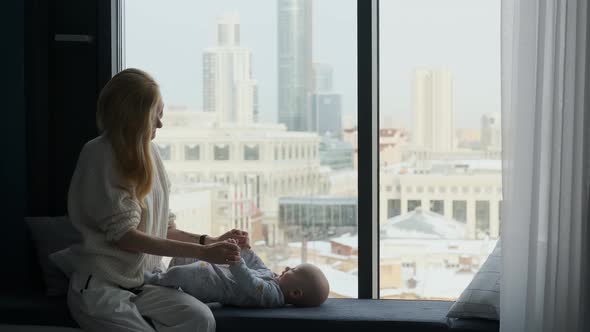 other doing gymnastics to her baby while sitting by the window with city view