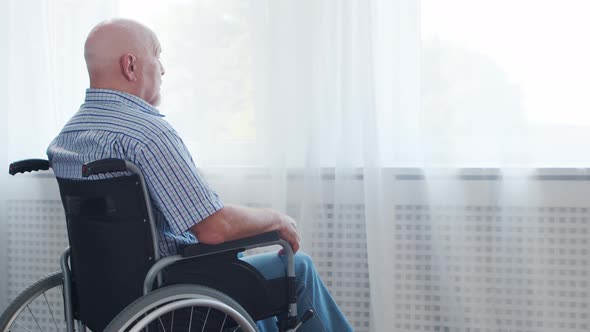 Disabled old man is sitting in a wheelchair at home alone. A handicapped person. alt