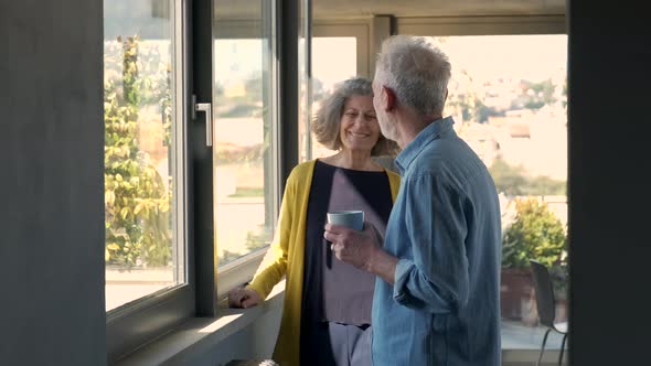 Senior woman embracing man looking out of window with coffee cup alt