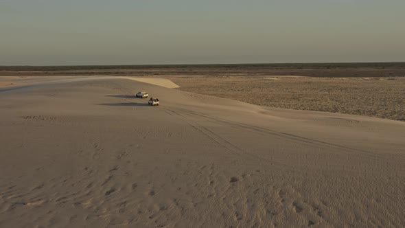Offroad vehicles explore windy Brazil dunes with sand gusts in sunset; drone alt