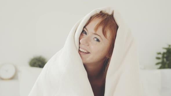 Smiling cheerful woman looks out from under the blanket in bed at home