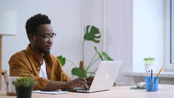 African Man in Glasses on Laptop Works in White Home Interior Background of Window Spbas alt