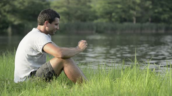 Young Man in Nature Seated on Grass Take Photo with Smartphone on Lake Shore in Sunny Summer Day