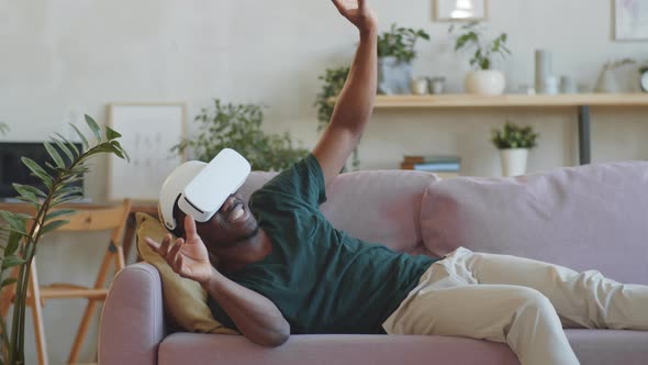 Excited Afro-American Man Lying on Sofa and Using VR Headset alt