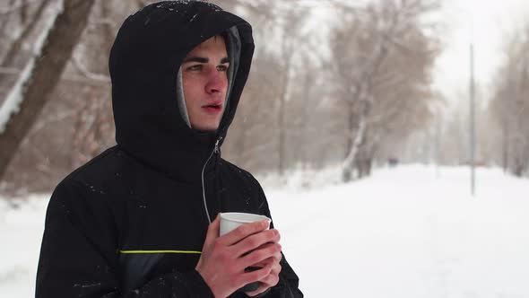 Caucasian man in black jacket with hood is warming himself in cold and drinking hot coffee. alt