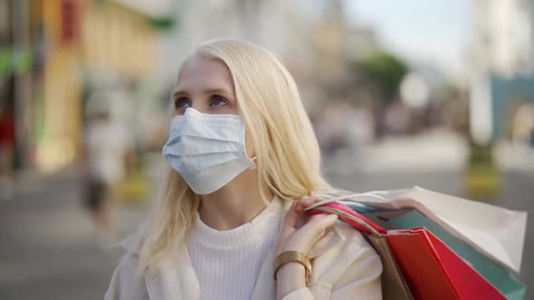 A Young Blonde Woman in a Medical Mask with Paktemi From Different Clothing Stores Walks Down the