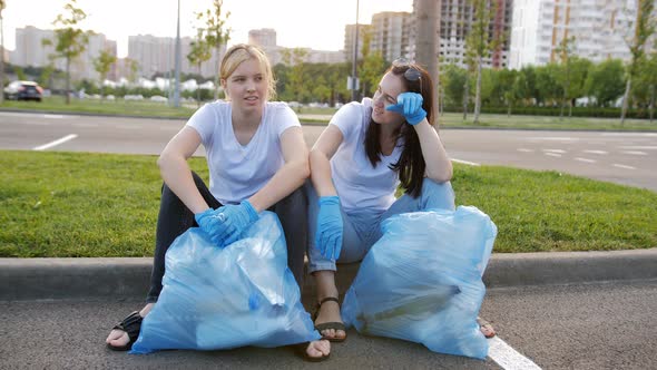 A Woman and a Teenage Girl are Sitting Together on the Curb with Large Blue Garbage Bags