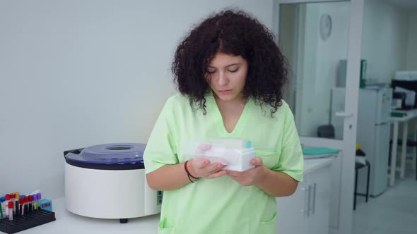 Concentrated Plussize Lab Assistant Examining Test Tubes Box and Looking at Camera Smiling alt
