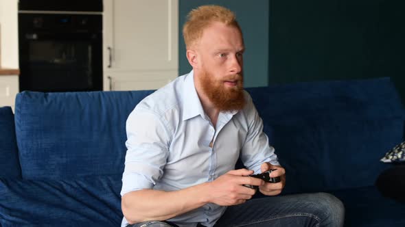 Upset Young Redhead Man Loses in Video Game Sitting on the Couch at Home with Joystick