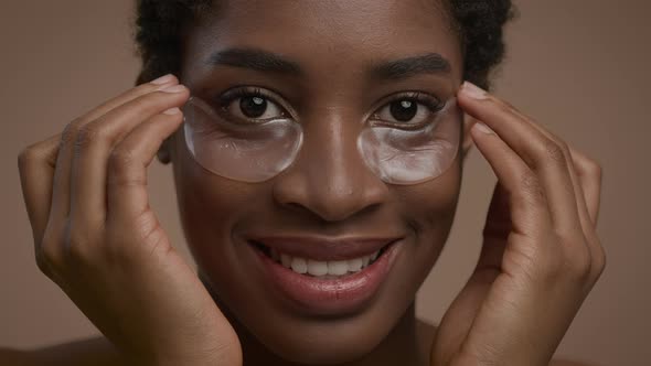Pretty African American Woman Applying UnderEye Patches Over Beige Background alt