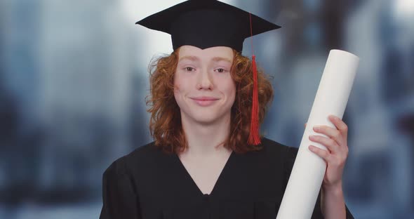 Young Student in a Robe and Hat Holds a Diploma Paper Scroll in His Hands, a Red-haired Teenager alt