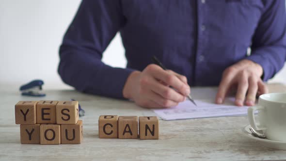 Motivating Message Yes You Can Solved Using Cubes. Man Works At Table, Checking Documents alt