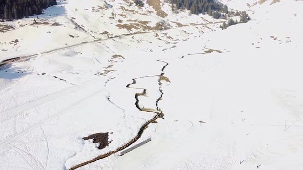 Drone shot ing forwards) of a Stream in the middle of a Snowy Mountain Plain with a Road Behind alt