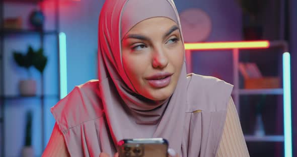 Muslim Woman in Hijab Sitting in Colourful Lighted Room in the Evening and Using smartphone alt