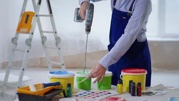 Male Construction Worker in Work Clothes and Helmet Mixes Paint in Bucket Using Drill To Paint Walls alt