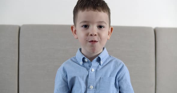 A Cute Little Boy Looks at the Camera and Sitting on the Couch at Home and Eats a Bar of Chocolate alt