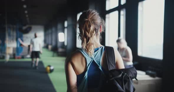 Camera Follows Young Athletic Blonde Woman with Backpack Entering Large Modern Gym Greeting Friends alt