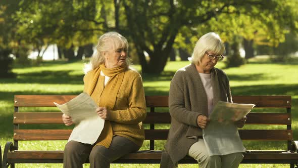 Two Senior Offended Female Friends Reading Newspapers, Sitting on Bench in Park alt