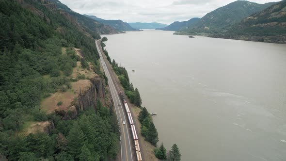 Aerial rise above train and highway traffic traveling next to Columbia River. alt